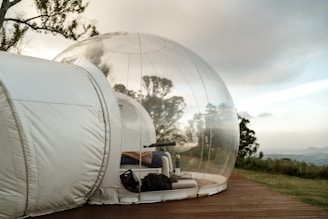 A transparent dome structure with a connected narrow extension stands on a wooden deck amidst a natural, serene landscape. Inside, a cozy setup includes a bed, a blanket, and a few personal items. Trees and a grassy field are visible in the background, along with a cloudy sky.