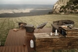 A serene outdoor setting featuring a wooden deck with lounge chairs and a couch. There is a fire pit with wood logs nearby, and a small table with items like food, a kettle, and bottles. The scene is surrounded by lush grass and overlooks a breathtaking mountainous landscape.