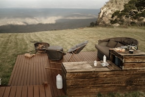 A serene outdoor setting featuring a wooden deck with lounge chairs and a couch. There is a fire pit with wood logs nearby, and a small table with items like food, a kettle, and bottles. The scene is surrounded by lush grass and overlooks a breathtaking mountainous landscape.