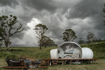 A modern, transparent bubble tent is set amidst an open, grassy field with a backdrop of dramatic, dark clouds and tall trees. The tent is placed on a wooden platform and is equipped with outdoor seating and a small firewood stack nearby, indicating a cozy outdoor setting.
