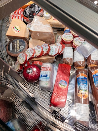 A variety of cheeses and cured meats displayed in a refrigerated glass case, featuring several kinds of cheese with labels in different colors and shapes. The assortment includes round, wedge-shaped, and cylindrical pieces, with visible branding and pricing information.