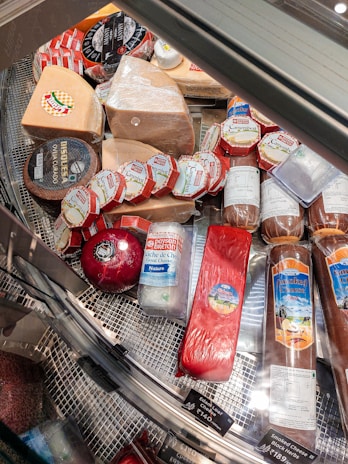 A variety of cheeses and cured meats displayed in a refrigerated glass case, featuring several kinds of cheese with labels in different colors and shapes. The assortment includes round, wedge-shaped, and cylindrical pieces, with visible branding and pricing information.
