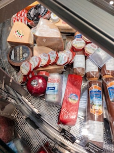A variety of cheeses and cured meats displayed in a refrigerated glass case, featuring several kinds of cheese with labels in different colors and shapes. The assortment includes round, wedge-shaped, and cylindrical pieces, with visible branding and pricing information.