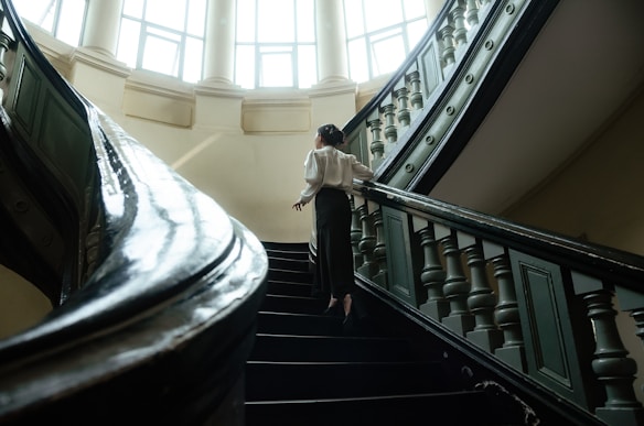 A woman in formal attire ascends a grand, curved staircase inside a well-lit, elegant building. The architecture features tall windows and intricately designed railings.