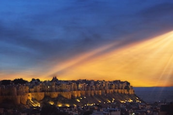 A majestic fort is perched atop a rocky hill, surrounded by a sprawling cityscape. The fort is illuminated by warm lights, casting a golden glow. The sky is painted with hues of deep blue and orange, with dramatic rays of sunlight piercing through clouds, creating a striking atmospheric effect.