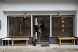 A storefront with a central glass door and two large windows on either side, featuring wooden benches in front. The windows have horizontal blinds and one of them has Chinese or Japanese characters. There are hanging lights above each window and the door, and a small table and potted plant are placed near the entrance.