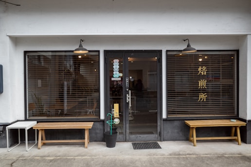 A storefront with a central glass door and two large windows on either side, featuring wooden benches in front. The windows have horizontal blinds and one of them has Chinese or Japanese characters. There are hanging lights above each window and the door, and a small table and potted plant are placed near the entrance.