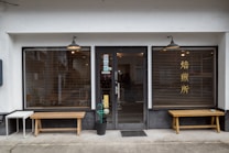 A storefront with a central glass door and two large windows on either side, featuring wooden benches in front. The windows have horizontal blinds and one of them has Chinese or Japanese characters. There are hanging lights above each window and the door, and a small table and potted plant are placed near the entrance.