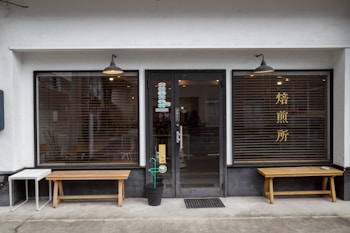 A storefront with a central glass door and two large windows on either side, featuring wooden benches in front. The windows have horizontal blinds and one of them has Chinese or Japanese characters. There are hanging lights above each window and the door, and a small table and potted plant are placed near the entrance.