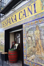The entrance to a Spanish establishment is decorated with colorful ceramic tiles. Above the door, large lettering spells out 'España Cañí' with intricate border designs. To the right, a vivid mural features historical or mythological scenes, including people and a guitar. A red barrel serves as a table near the entrance, adding to the rustic ambiance.