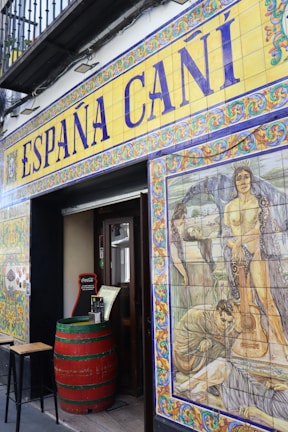 The entrance to a Spanish establishment is decorated with colorful ceramic tiles. Above the door, large lettering spells out 'España Cañí' with intricate border designs. To the right, a vivid mural features historical or mythological scenes, including people and a guitar. A red barrel serves as a table near the entrance, adding to the rustic ambiance.