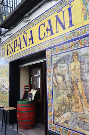 The entrance to a Spanish establishment is decorated with colorful ceramic tiles. Above the door, large lettering spells out 'Espa&ntilde;a Ca&ntilde;&iacute;' with intricate border designs. To the right, a vivid mural features historical or mythological scenes, including people and a guitar. A red barrel serves as a table near the entrance, adding to the rustic ambiance.