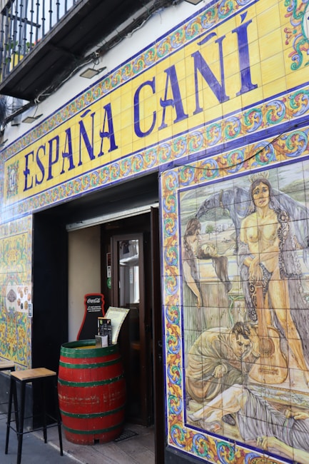 The entrance to a Spanish establishment is decorated with colorful ceramic tiles. Above the door, large lettering spells out 'Espa&ntilde;a Ca&ntilde;&iacute;' with intricate border designs. To the right, a vivid mural features historical or mythological scenes, including people and a guitar. A red barrel serves as a table near the entrance, adding to the rustic ambiance.