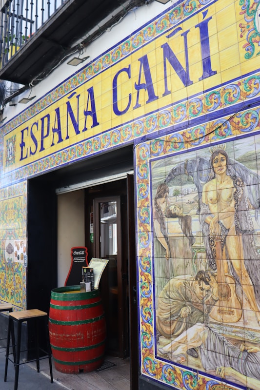 The entrance to a Spanish establishment is decorated with colorful ceramic tiles. Above the door, large lettering spells out 'Espa&ntilde;a Ca&ntilde;&iacute;' with intricate border designs. To the right, a vivid mural features historical or mythological scenes, including people and a guitar. A red barrel serves as a table near the entrance, adding to the rustic ambiance.