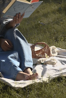 A person is sitting on a white blanket spread on the grass, reading a light blue book titled 'IKIGAI'. The person is wearing denim jeans and rings on their fingers. Beside them, there is a small, woven basket, and sunlight casts soft shadows on the scene.