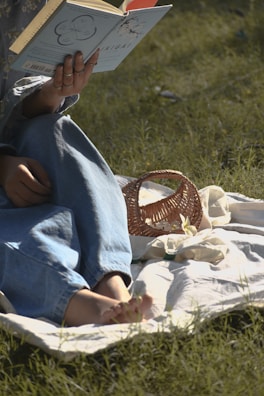 A person is sitting on a white blanket spread on the grass, reading a light blue book titled 'IKIGAI'. The person is wearing denim jeans and rings on their fingers. Beside them, there is a small, woven basket, and sunlight casts soft shadows on the scene.