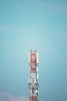 A communication tower with various antennas and equipment attached to it stands against a clear blue sky. The structure is primarily composed of metal and painted in red and white. Cables and wires are visible running along the tower.