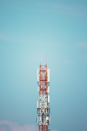 A communication tower with various antennas and equipment attached to it stands against a clear blue sky. The structure is primarily composed of metal and painted in red and white. Cables and wires are visible running along the tower.
