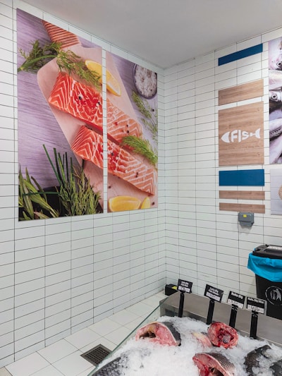 A brightly lit fish market section featuring a tiled wall with large posters of raw salmon fillets garnished with herbs and lemon slices. Below, a display of various fish cuts on ice with labeled price tags. The corner extends to another wall decorated with signs and images related to fish.