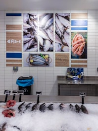 A display of fresh seafood in a market, including several whole fish and fish heads on ice, with price tags indicating their types and costs. The wall behind features decorative images of fish and shrimp, along with nautical-themed artwork and signage.