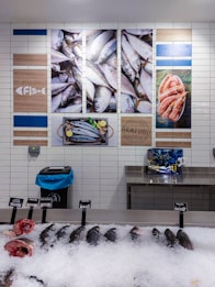 A display of fresh seafood in a market, including several whole fish and fish heads on ice, with price tags indicating their types and costs. The wall behind features decorative images of fish and shrimp, along with nautical-themed artwork and signage.