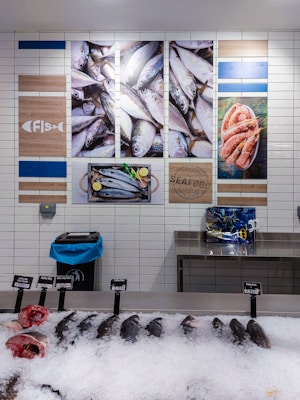 A display of fresh seafood in a market, including several whole fish and fish heads on ice, with price tags indicating their types and costs. The wall behind features decorative images of fish and shrimp, along with nautical-themed artwork and signage.