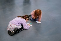 Two children are kneeling on the floor, engrossed in drawing or writing on pieces of paper. Both are wearing colorful and cozy clothing, with one child in a purple sweater and the other in pink. The setting appears to be a smooth, open indoor area.