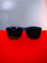 A pair of dark sunglasses rests on a vibrant red surface, with a smooth grayish backdrop creating a modern and minimalistic look. The reflection on the sunglasses adds a subtle touch of depth to the composition.
