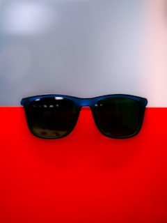 A pair of dark sunglasses rests on a vibrant red surface, with a smooth grayish backdrop creating a modern and minimalistic look. The reflection on the sunglasses adds a subtle touch of depth to the composition.