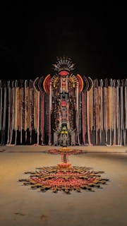 A large, intricate installation made of various materials hangs against a dark background. It features symmetrical patterns and uses a combination of colors including reds, golds, and blacks, arranged in a way that resembles a tribal or ceremonial design. Long strands cascade from the central structure, and a geometric pattern extends on the floor below.