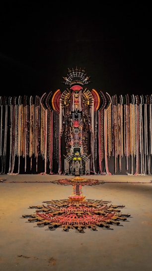A large, intricate installation made of various materials hangs against a dark background. It features symmetrical patterns and uses a combination of colors including reds, golds, and blacks, arranged in a way that resembles a tribal or ceremonial design. Long strands cascade from the central structure, and a geometric pattern extends on the floor below.