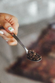 A hand holds a spoonful of coffee beans with a soft focus background. The person's nails are painted white with a small green avocado design on one nail.