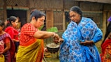 Two women are engaging in a traditional activity, with one woman wearing a red and yellow saree passing something from a bowl to a woman in a blue and white shawl. Several other women are present in the background, wearing colorful traditional garments. The setting appears to be outdoors, near a building with wooden and bamboo features.