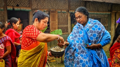 Two women are engaging in a traditional activity, with one woman wearing a red and yellow saree passing something from a bowl to a woman in a blue and white shawl. Several other women are present in the background, wearing colorful traditional garments. The setting appears to be outdoors, near a building with wooden and bamboo features.