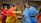 Two women are engaging in a traditional activity, with one woman wearing a red and yellow saree passing something from a bowl to a woman in a blue and white shawl. Several other women are present in the background, wearing colorful traditional garments. The setting appears to be outdoors, near a building with wooden and bamboo features.