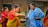 Two women are engaging in a traditional activity, with one woman wearing a red and yellow saree passing something from a bowl to a woman in a blue and white shawl. Several other women are present in the background, wearing colorful traditional garments. The setting appears to be outdoors, near a building with wooden and bamboo features.
