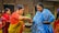 Two women are engaging in a traditional activity, with one woman wearing a red and yellow saree passing something from a bowl to a woman in a blue and white shawl. Several other women are present in the background, wearing colorful traditional garments. The setting appears to be outdoors, near a building with wooden and bamboo features.