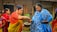 Two women are engaging in a traditional activity, with one woman wearing a red and yellow saree passing something from a bowl to a woman in a blue and white shawl. Several other women are present in the background, wearing colorful traditional garments. The setting appears to be outdoors, near a building with wooden and bamboo features.