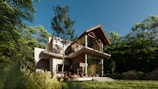 A modern two-story house with large glass windows is surrounded by dense forest. The building features a sloped wooden roof and a spacious balcony. The lush greenery adds to the serene atmosphere with sunlight filtering through the trees onto the grass.