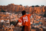 A young boy with outstretched arms stands against a backdrop of an ancient or rustic cityscape. He is wearing a bright red shirt with the word 'HOPE' printed on the back. The background is composed of earthy, sandy-colored, traditional buildings, likely portraying a historical or old town setting.