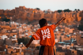 A young boy with outstretched arms stands against a backdrop of an ancient or rustic cityscape. He is wearing a bright red shirt with the word 'HOPE' printed on the back. The background is composed of earthy, sandy-colored, traditional buildings, likely portraying a historical or old town setting.