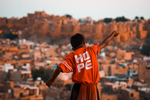 A young boy with outstretched arms stands against a backdrop of an ancient or rustic cityscape. He is wearing a bright red shirt with the word 'HOPE' printed on the back. The background is composed of earthy, sandy-colored, traditional buildings, likely portraying a historical or old town setting.