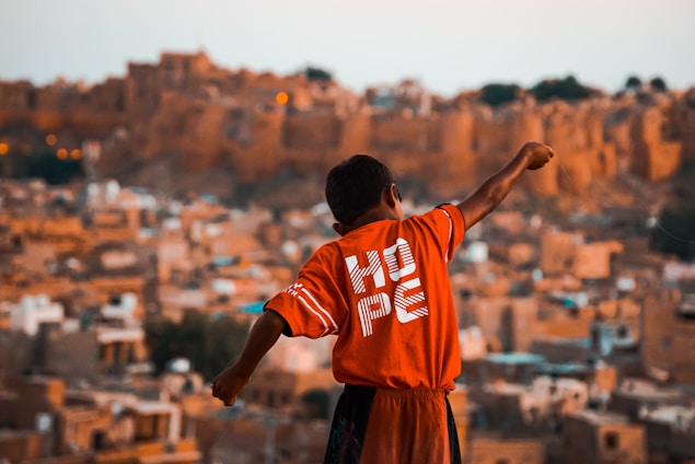 A young boy with outstretched arms stands against a backdrop of an ancient or rustic cityscape. He is wearing a bright red shirt with the word 'HOPE' printed on the back. The background is composed of earthy, sandy-colored, traditional buildings, likely portraying a historical or old town setting.