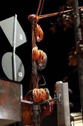 A utility pole is wrapped with several coils of thick orange cables. The cables are organized and bundled around the pole with gray clips. There are attached signs and a traffic light visible, suggesting it's an urban setup. The background is a dark night sky with some leafless tree branches partially visible.