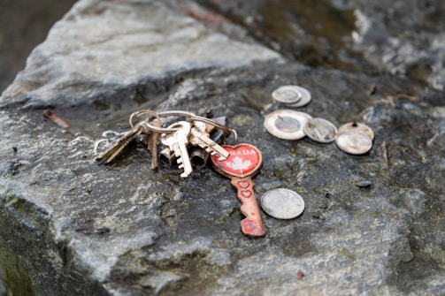 A collection of various keys on a ring and a few coins lying on a rough, textured stone surface. One of the keys has a distinctive red design with a maple leaf and the word 'Canada' on it, along with heart-shaped cutouts.
