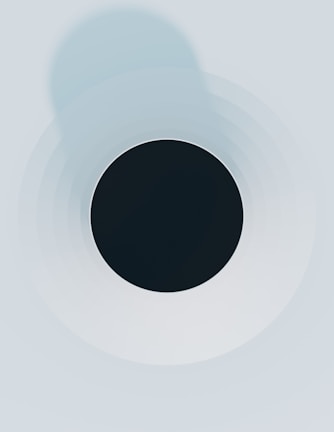 A minimalist abstract design featuring a black circle in the center surrounded by concentric circles that gradually fade into lighter shades of gray and blue. The composition has a clean and symmetrical appearance.
