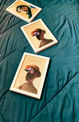 Three framed artworks placed on a dark green quilted surface. The artwork features stylized portraits with a detective theme, including illustrations of figures wearing hats and glasses, with one frame titled 'DETECTIVE'. The frames are positioned diagonally across the surface.