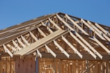 A detailed view of a wooden structure framework under construction, showcasing intersecting beams and trusses with a clear blue sky in the background.