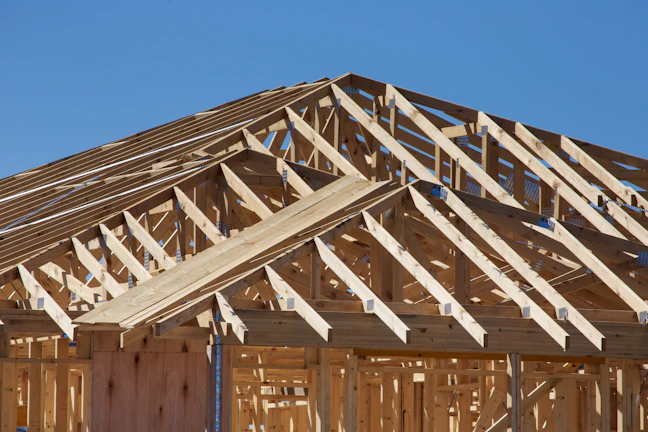 A detailed view of a wooden structure framework under construction, showcasing intersecting beams and trusses with a clear blue sky in the background.