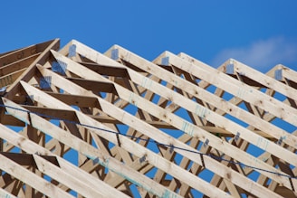 Wooden roof trusses arranged in a triangular pattern against a clear blue sky. Metal connectors hold the beams together, indicating the early stage of construction.